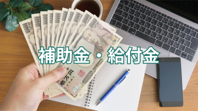 パソコン・ノート・スマホ・ペンが置かれたデスク上で一万円札を数枚広げて持っている様子「補助金・給付金」