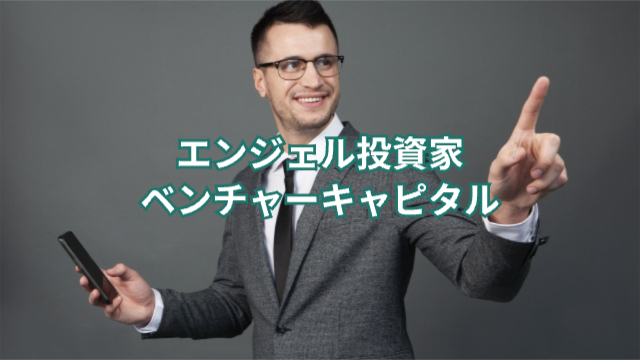 投資家の男性がスマホを持って笑顔で人差し指を立てている「エンジェル投資家・ベンチャーキャピタル」