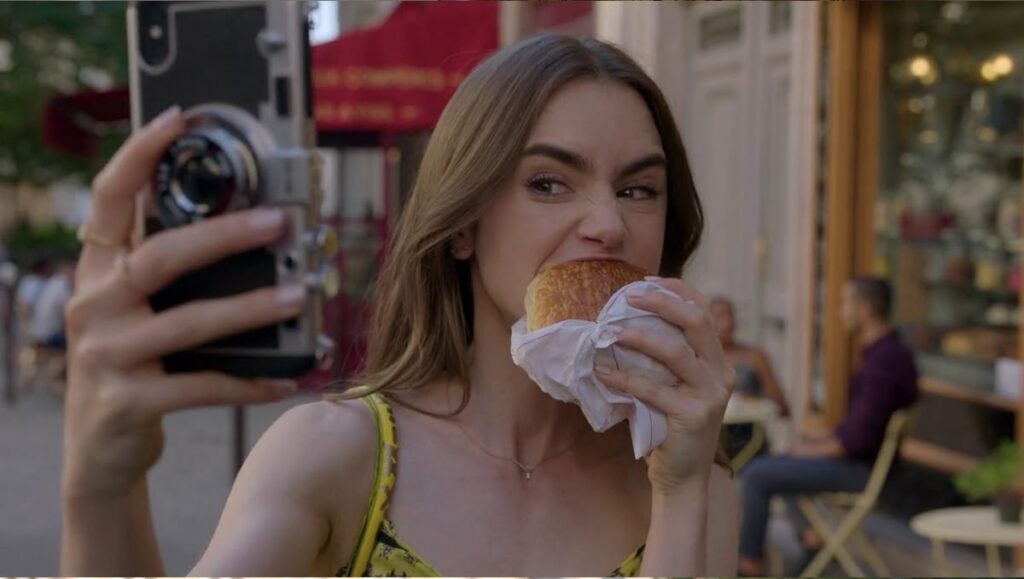 エミリー、パリへ行くのクロワッサンを食べて自撮りしているシーン
